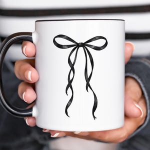 May include: White ceramic mug with a black interior and handle. The mug features a black bow design on the front. The bow has long, flowing ribbons. The mug is held by a person.