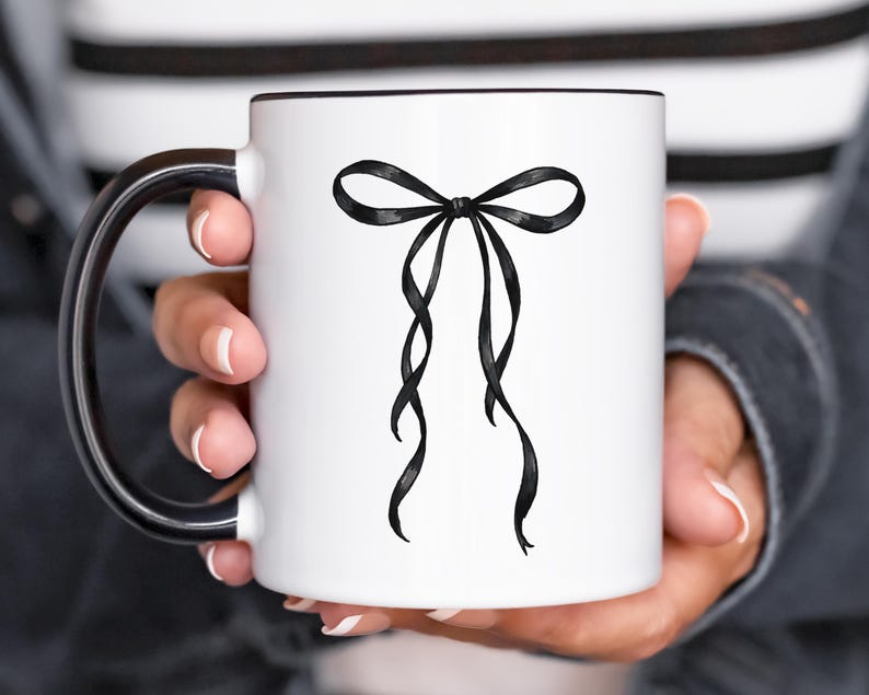 May include: White ceramic mug with a black interior and handle. The mug features a black bow design on the front. The bow has long, flowing ribbons. The mug is held by a person.