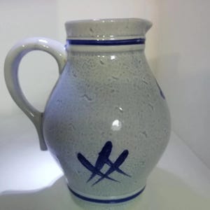 Peut inclure: Une cruche en céramique gris clair avec une texture mouchetée, dotée d'une anse incurvée et d'un bec verseur. Le récipient est orné de bandes bleues horizontales autour du col et de la base, ainsi qu'une marque abstraite peinte en bleu.