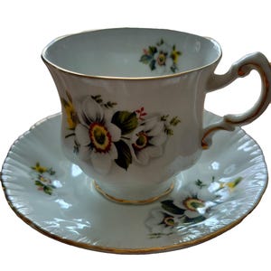 Peut inclure: Un ensemble tasse et soucoupe en porcelaine blanche orné d'un motif floral délicat de fleurs blanches aux centres jaunes et rouges. La tasse présente un bord festonné et une anse incurvée, tous deux rehaussés d'un liseré doré assorti.