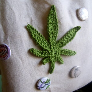 May include: A handmade, green crochet cannabis leaf is centered on a cream-colored fabric. Three decorative pinback buttons are scattered around the leaf. The buttons have various designs and text, including the word "spin" and "garden crochet".