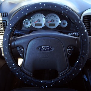 May include: A black steering wheel cover with white stars. The steering wheel has a black leather surface and a silver Ford logo.