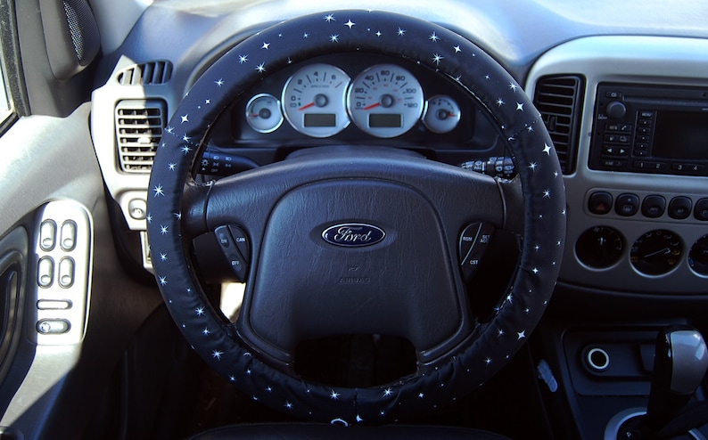 May include: A black steering wheel cover with white stars. The steering wheel has a black leather surface and a silver Ford logo.