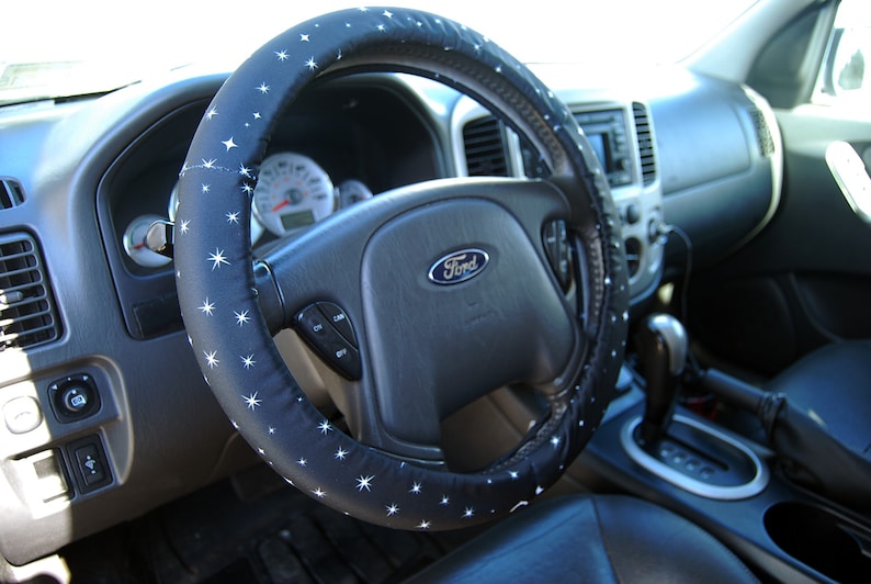 May include: A black steering wheel cover with white stars. The cover is installed on a black leather steering wheel with a silver Ford logo.