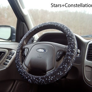 May include: A black steering wheel cover with a white star and constellation pattern. The cover is installed on a car's steering wheel.