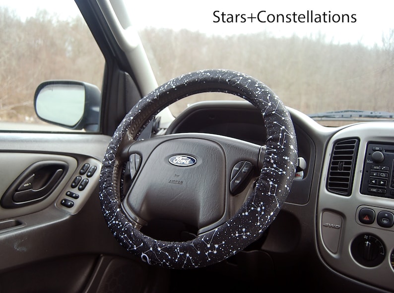 May include: A black steering wheel cover with a white star and constellation pattern. The cover is installed on a car's steering wheel.