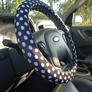 Pu&ograve; includere: Un coprivolante nero con pois bianchi &egrave; installato sul volante di un'auto. L'interno dell'auto &egrave; visibile, compreso il cruscotto, i sedili e la portiera.
