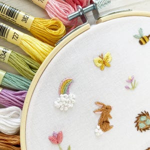 Op de afbeelding: Een close-up van een witte borduurring met verschillende kleurrijke borduurmotieven, waaronder een regenboog, een konijn, een vlinder en een bij. De borduurring is omgeven door verschillende borduurgaren in verschillende kleuren.