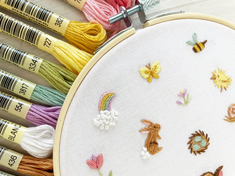 Op de afbeelding: Een close-up van een witte borduurring met verschillende kleurrijke borduurmotieven, waaronder een regenboog, een konijn, een vlinder en een bij. De borduurring is omgeven door verschillende borduurgaren in verschillende kleuren.
