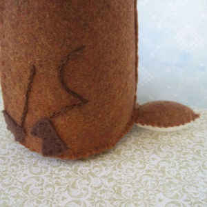 May include: A brown felt toy with a long tail and a stitched design on the back. The toy is sitting on a white and gold patterned surface.