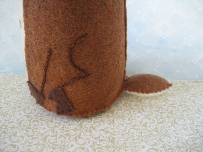 May include: A brown felt toy with a long tail and a stitched design on the back. The toy is sitting on a white and gold patterned surface.