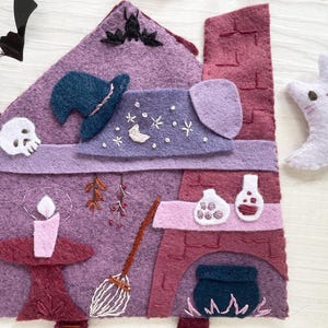 May include: A felt Halloween-themed house in shades of purple and burgundy, with a witch's hat, skull, and a crescent moon with a cat face. The house features a candle, broom, and a fireplace with a cauldron.