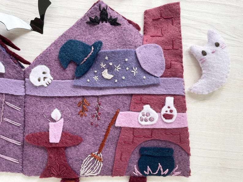 May include: A felt Halloween-themed house in shades of purple and burgundy, with a witch's hat, skull, and a crescent moon with a cat face. The house features a candle, broom, and a fireplace with a cauldron.