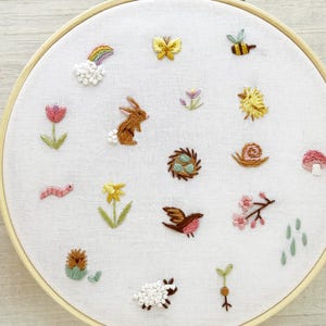Op de afbeelding: Een witte borduurring met verschillende kleurrijke geborduurde ontwerpen, waaronder een regenboog, een vlinder, een konijn, een vogel, een slak, een paddenstoel, een bloem, een worm, een egel, een schaap en een scheut.