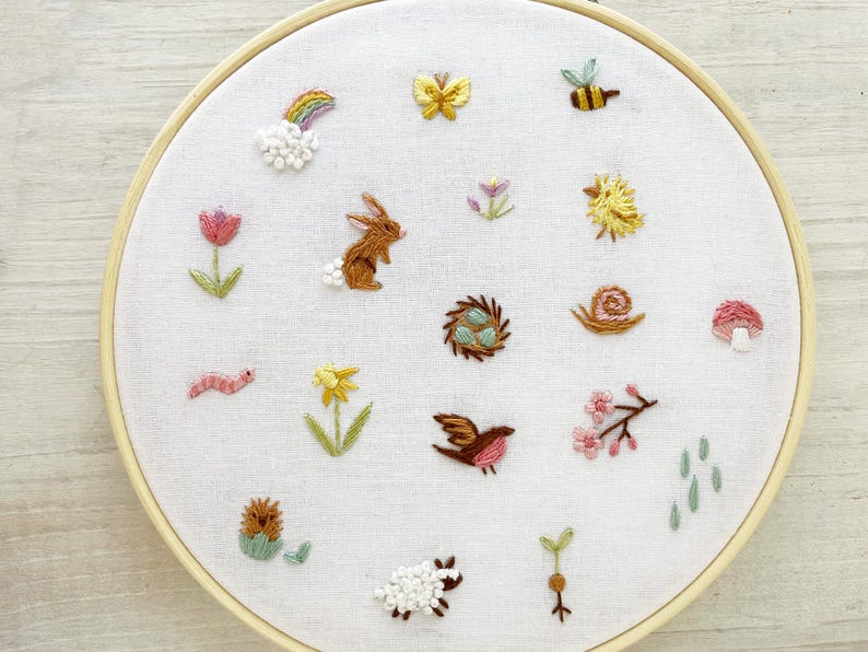 Op de afbeelding: Een witte borduurring met verschillende kleurrijke geborduurde ontwerpen, waaronder een regenboog, een vlinder, een konijn, een vogel, een slak, een paddenstoel, een bloem, een worm, een egel, een schaap en een scheut.