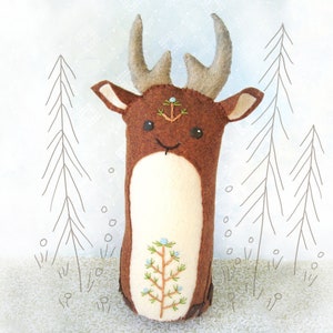 May include: A brown felt deer with a white belly and embroidered details. The deer has large antlers and a small pine tree embroidered on its forehead. The deer is standing in front of a white background with a drawing of pine trees.