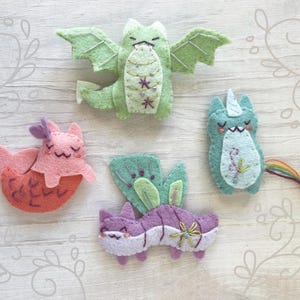 May include: Five felt animal ornaments, including a green dragon, a pink mermaid, a blue unicorn, a purple butterfly, and a green and purple cat. The ornaments are all hand-stitched and have embroidered details.