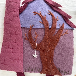 May include: A felt Halloween decoration featuring a purple house with a brown tree and a purple roof. A white star hangs from the tree. A dark purple chimney is on the left. A black bat and star decorations are in the background.