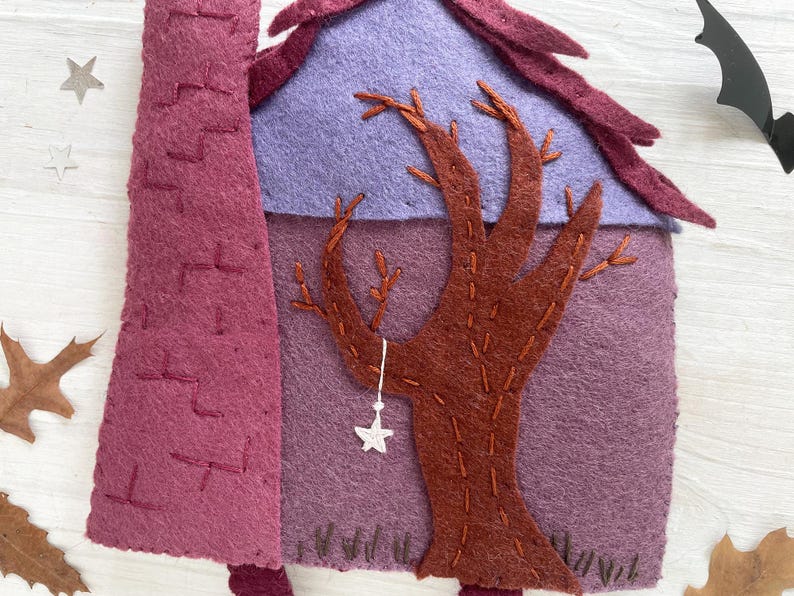 May include: A felt Halloween decoration featuring a purple house with a brown tree and a purple roof. A white star hangs from the tree. A dark purple chimney is on the left. A black bat and star decorations are in the background.