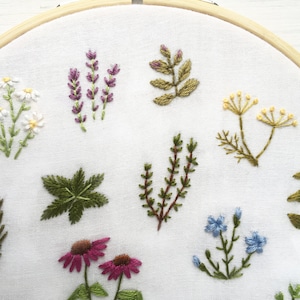 May include: Embroidered hoop art featuring a variety of flowers and plants, including lavender, chamomile, and ferns. The embroidery is done in shades of green, purple, white, and yellow on a white fabric.