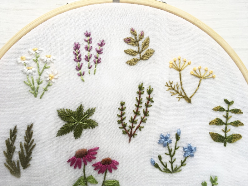 May include: Embroidered hoop art featuring a variety of flowers and plants, including lavender, chamomile, and ferns. The embroidery is done in shades of green, purple, white, and yellow on a white fabric.