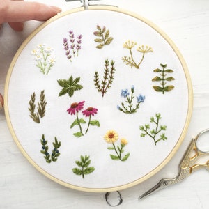 May include: A white embroidery hoop with a variety of green, yellow, pink, and blue embroidered flowers and leaves. The hoop is on a white surface with a pair of gold and silver embroidery scissors.