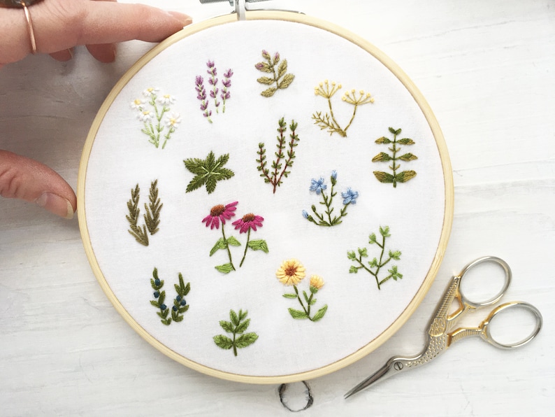 May include: A white embroidery hoop with a variety of green, yellow, pink, and blue embroidered flowers and leaves. The hoop is on a white surface with a pair of gold and silver embroidery scissors.