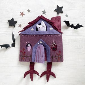 May include: A whimsical felt house in shades of purple, with a cat peering from the window. The house has a red roof, a purple door, and is decorated with stars and a candle. Black bat and star cutouts surround the house.