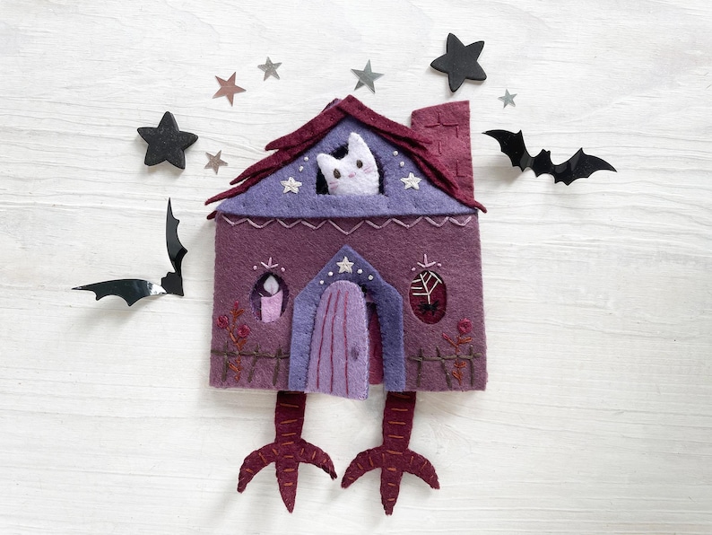 May include: A whimsical felt house in shades of purple, with a cat peering from the window. The house has a red roof, a purple door, and is decorated with stars and a candle. Black bat and star cutouts surround the house.
