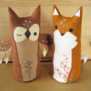 May include: Two felt animal plushies, an owl and a fox, with embroidered details. The owl is brown with white eyes and a light blue tree design. The fox is orange with white fur and a light pink floral design.
