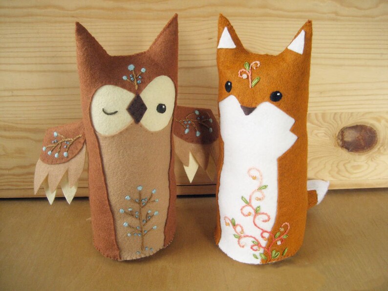 May include: Two felt animal plushies, an owl and a fox, with embroidered details. The owl is brown with white eyes and a light blue tree design. The fox is orange with white fur and a light pink floral design.