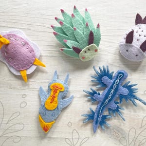 May include: A collection of five felt sea slug ornaments in various colors. The ornaments include purple, blue, and green sea slugs with yellow, red, and white accents. The felt ornaments are arranged on a light wood-grain surface.