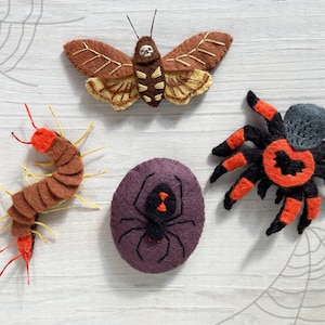 May include: A collection of handmade felt Halloween decorations. Includes a brown and tan moth, a brown centipede with red accents, a purple spider, and a black and orange spider. The decorations are arranged on a white wooden surface.
