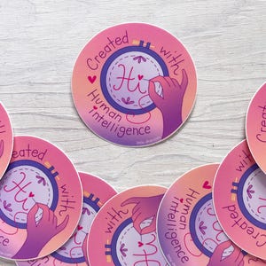 May include: A collection of round stickers with a pink and orange gradient background. Each sticker features a hand making the 'Hi' sign inside a cross-stitch hoop, with the text "Created with Human Intelligence".