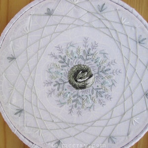 May include: White embroidered hoop art featuring a central nest design surrounded by delicate, light blue and gray leaves. The design is framed by a geometric pattern of intersecting lines, all on a white fabric background.