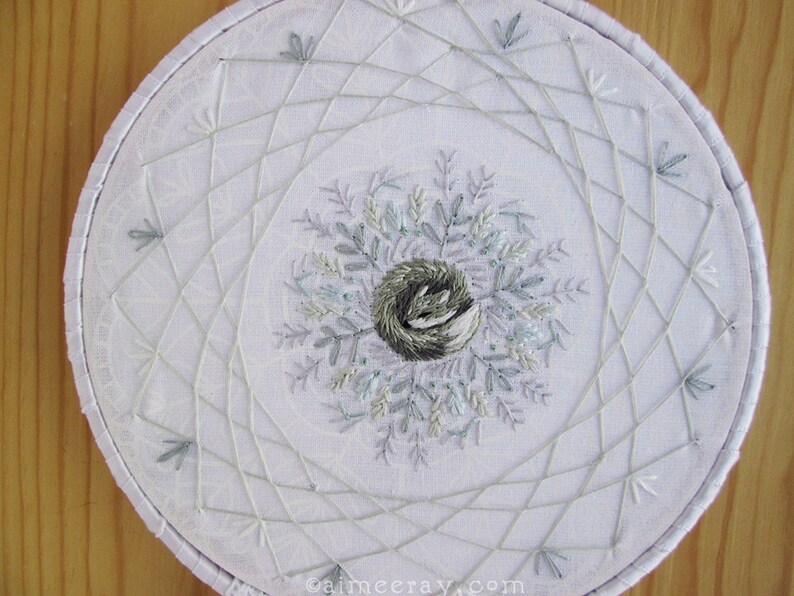 May include: White embroidered hoop art featuring a central nest design surrounded by delicate, light blue and gray leaves. The design is framed by a geometric pattern of intersecting lines, all on a white fabric background.