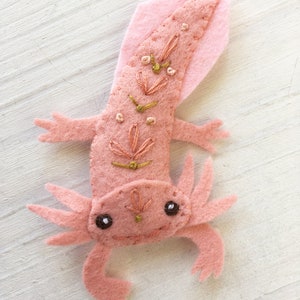 Pu&ograve; includere: Un axolotl di feltro rosa con dettagli ricamati, tra cui un motivo floreale rosa e oro sulla schiena e occhi neri. L'axolotl &egrave; un animale domestico popolare e un simbolo di dolcezza.