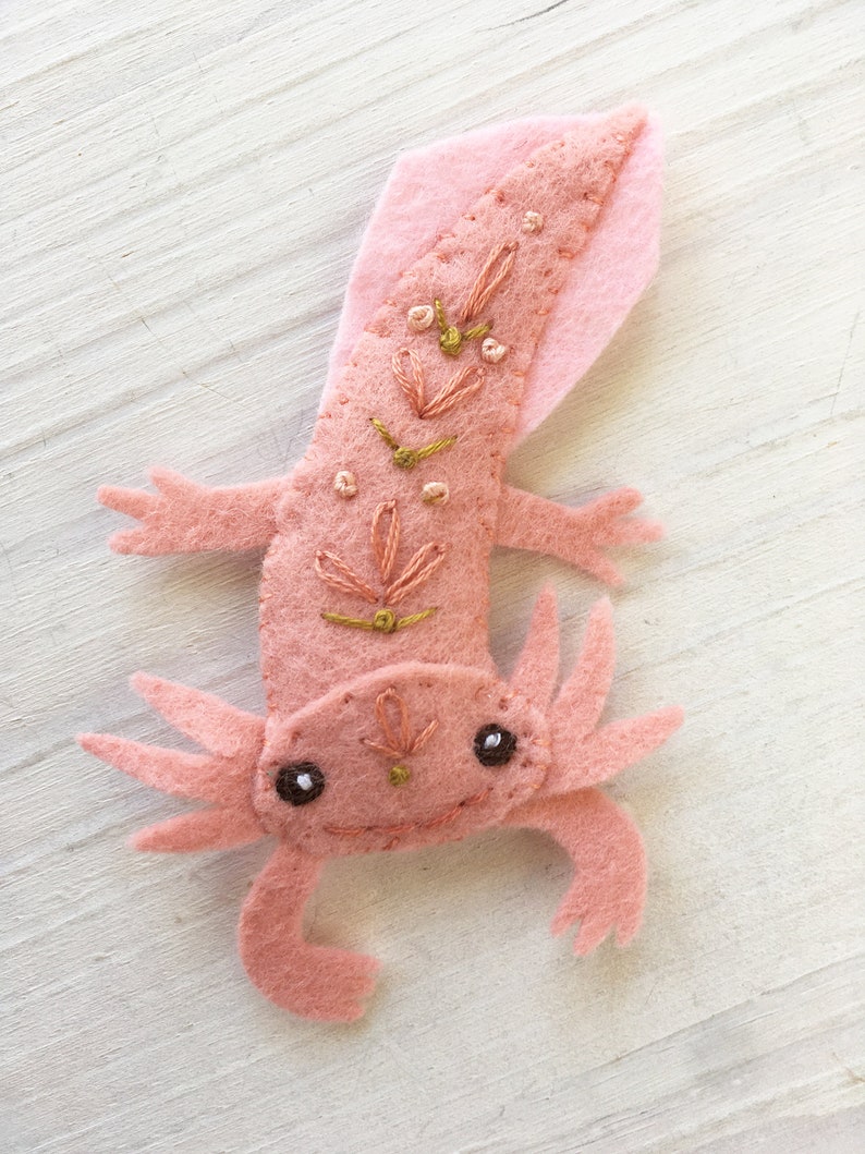 Pu&ograve; includere: Un axolotl di feltro rosa con dettagli ricamati, tra cui un motivo floreale rosa e oro sulla schiena e occhi neri. L'axolotl &egrave; un animale domestico popolare e un simbolo di dolcezza.