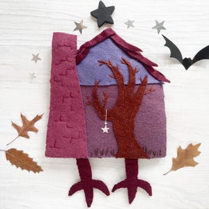 May include: A felt Halloween house with a chimney, tree, and star accents. The house is purple and burgundy, with a black star on top and a bat silhouette. Autumn leaves are scattered around the house.