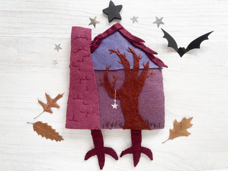 May include: A felt Halloween house with a chimney, tree, and star accents. The house is purple and burgundy, with a black star on top and a bat silhouette. Autumn leaves are scattered around the house.