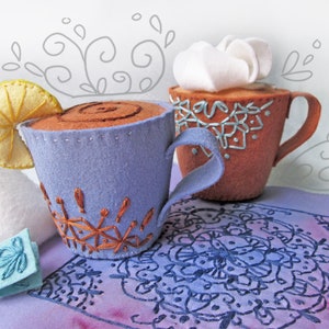 Puede incluir: Dos tazas de té de fieltro con detalles bordados. Una taza es morada con un diseño marrón y una rodaja de limón. La otra taza es marrón con un diseño blanco y un remolino blanco en la parte superior. Las tazas están sobre una tela morada con un diseño de mandala.