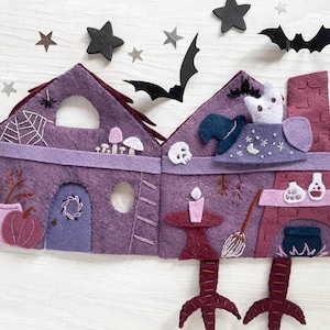 May include: A felt Halloween-themed house in shades of purple, with a witch's hat, a cat, and a cauldron. The house has a spiderweb, a pumpkin, and a broom. Black star and bat cutouts are scattered around the house.