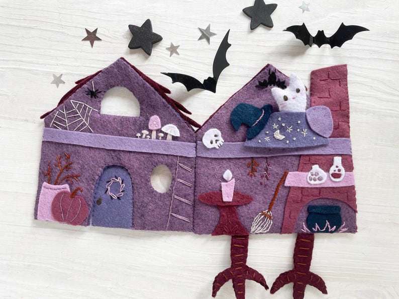 May include: A felt Halloween-themed house in shades of purple, with a witch's hat, a cat, and a cauldron. The house has a spiderweb, a pumpkin, and a broom. Black star and bat cutouts are scattered around the house.