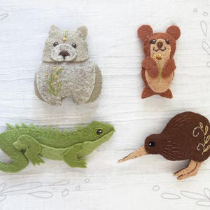 Puede incluir: Cuatro figuras de animales de fieltro: un oso gris, un oso marrón con una flor, un lagarto verde y un pájaro kiwi marrón. Cada figura tiene detalles bordados y está sobre un fondo blanco con un diseño floral.