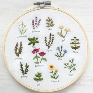 May include: A white embroidery hoop with a variety of embroidered herbs and flowers, including lavender, sage, dill, rosemary, mint, thyme, nettle, flax, echinacea, calendula, parsley, bilberry, and basil. Each plant is labeled with its name.