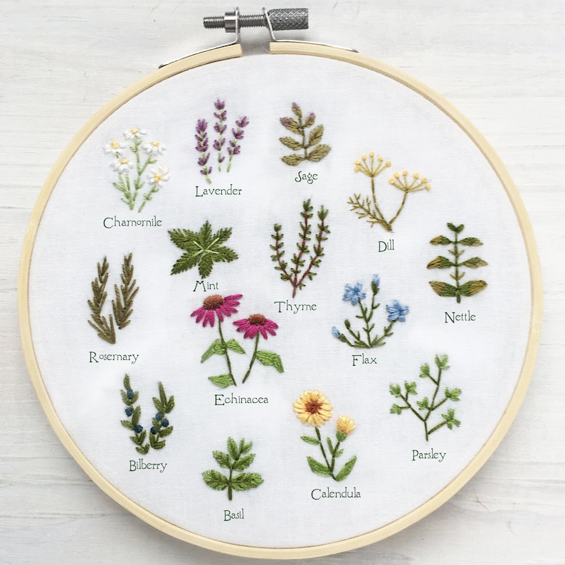 May include: A white embroidery hoop with a variety of embroidered herbs and flowers, including lavender, sage, dill, rosemary, mint, thyme, nettle, flax, echinacea, calendula, parsley, bilberry, and basil. Each plant is labeled with its name.