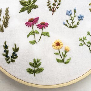 May include: Embroidered hoop art featuring a variety of floral and foliage designs. The hoop is made of wood and has a white fabric background. The embroidery includes pink coneflowers, yellow flowers, blue forget-me-nots, and green leaves.