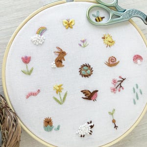 Op de afbeelding: Een witte borduurring met verschillende kleurrijke geborduurde ontwerpen, waaronder een konijn, een slak, een vogel, een bloem, een regenboog en een nest met eieren. De ring ligt op een wit houten oppervlak.