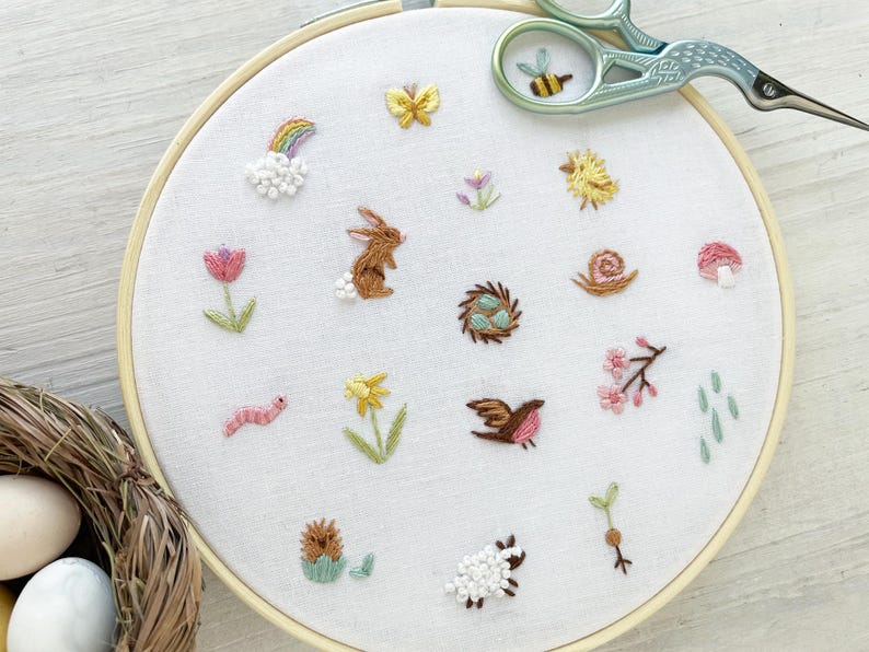Op de afbeelding: Een witte borduurring met verschillende kleurrijke geborduurde ontwerpen, waaronder een konijn, een slak, een vogel, een bloem, een regenboog en een nest met eieren. De ring ligt op een wit houten oppervlak.
