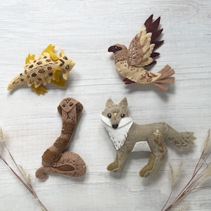 May include: Felt animal figures including a lizard, bird, snake, and fox. The lizard is yellow and brown, the bird is tan and brown, the snake is brown, and the fox is gray and white. The figures are arranged on a white wooden surface.